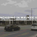 Street-level view of Kettlestone Lakes in Waukee, Iowa with retail storefronts, roadway traffic, landscaped commercial parcels, and franchise opportunities near I-80 and Grand Prairie Parkway.