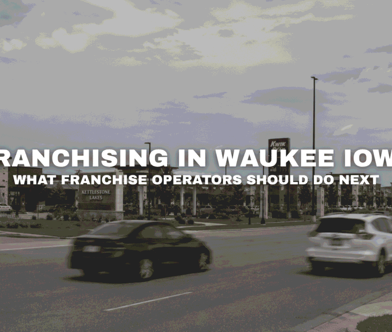 Street-level view of Kettlestone Lakes in Waukee, Iowa with retail storefronts, roadway traffic, landscaped commercial parcels, and franchise opportunities near I-80 and Grand Prairie Parkway.